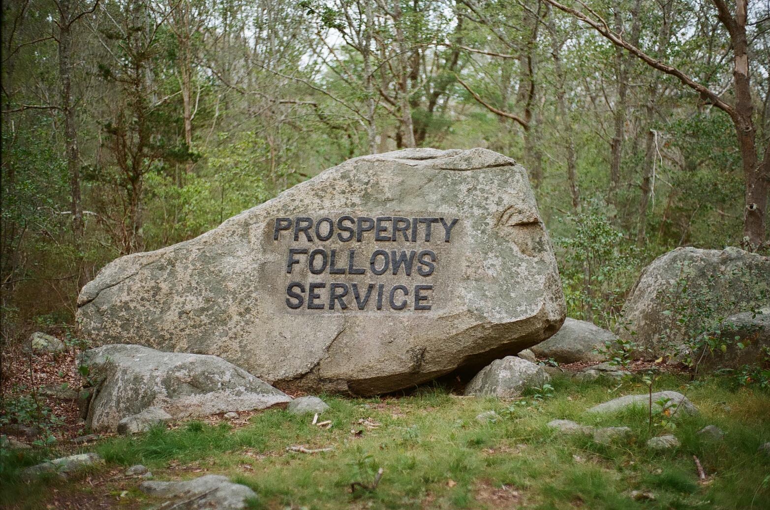 A boulder in the woods carved with the phrase: Prosperity Follows Service