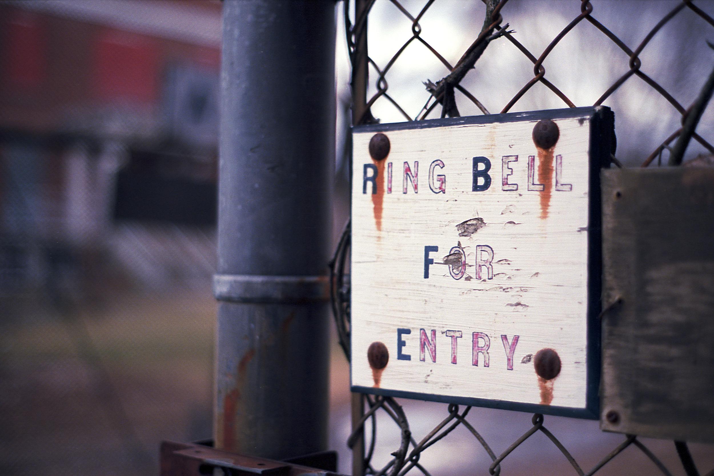 A rusted sign on a chain link fence that says Ring Bell For Entry