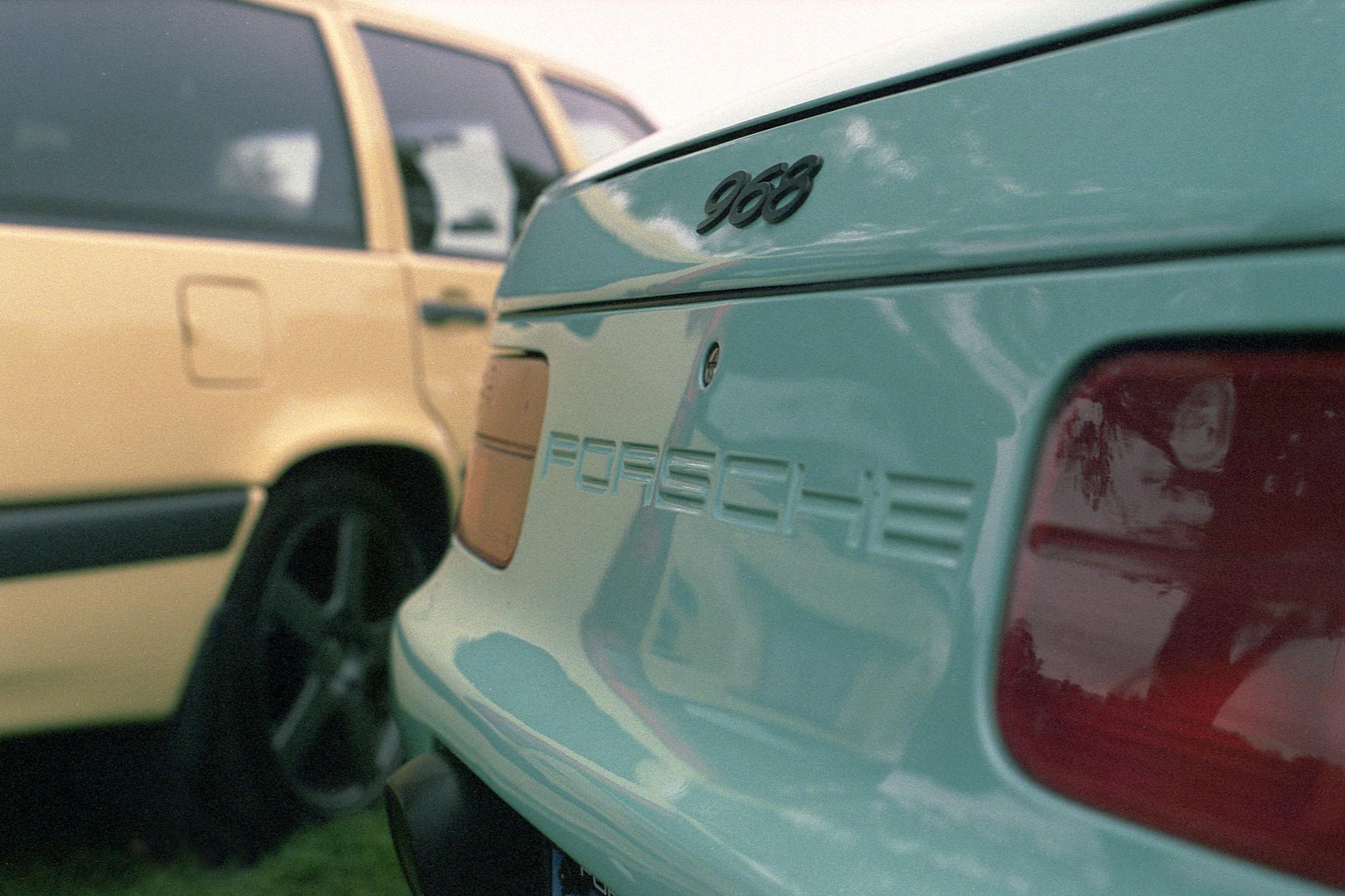 The rear of a turqoise Porsche 968 convertible in front of a yellow Volvo station wagon