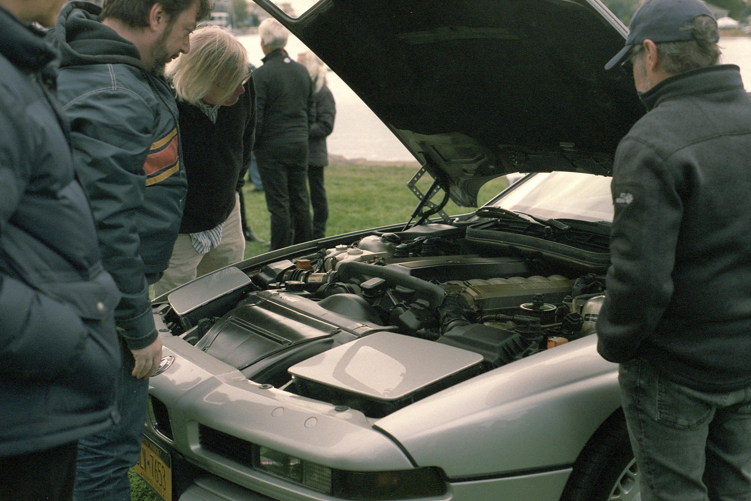 A group of people gathered around the open engine bay of an BMW E31 8 series