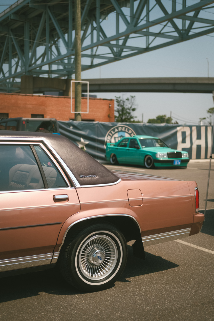 The rear end of a light brown Ford Crown Victoria with a teal Mercedes Benz in the distance