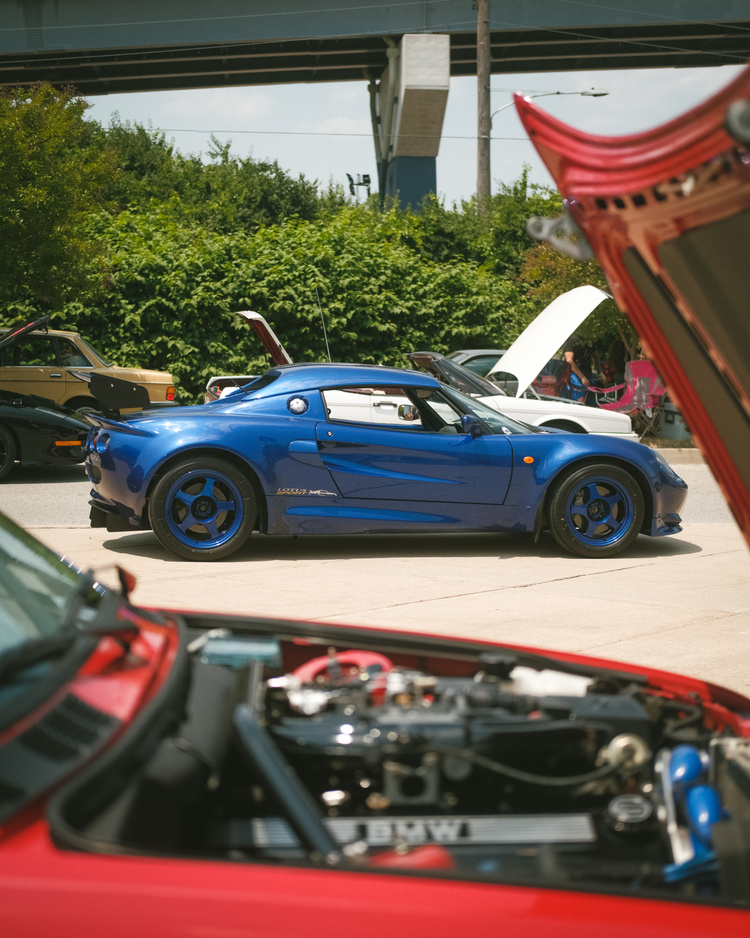 A blue Lotus Elise seen through the open hood of a red BMW