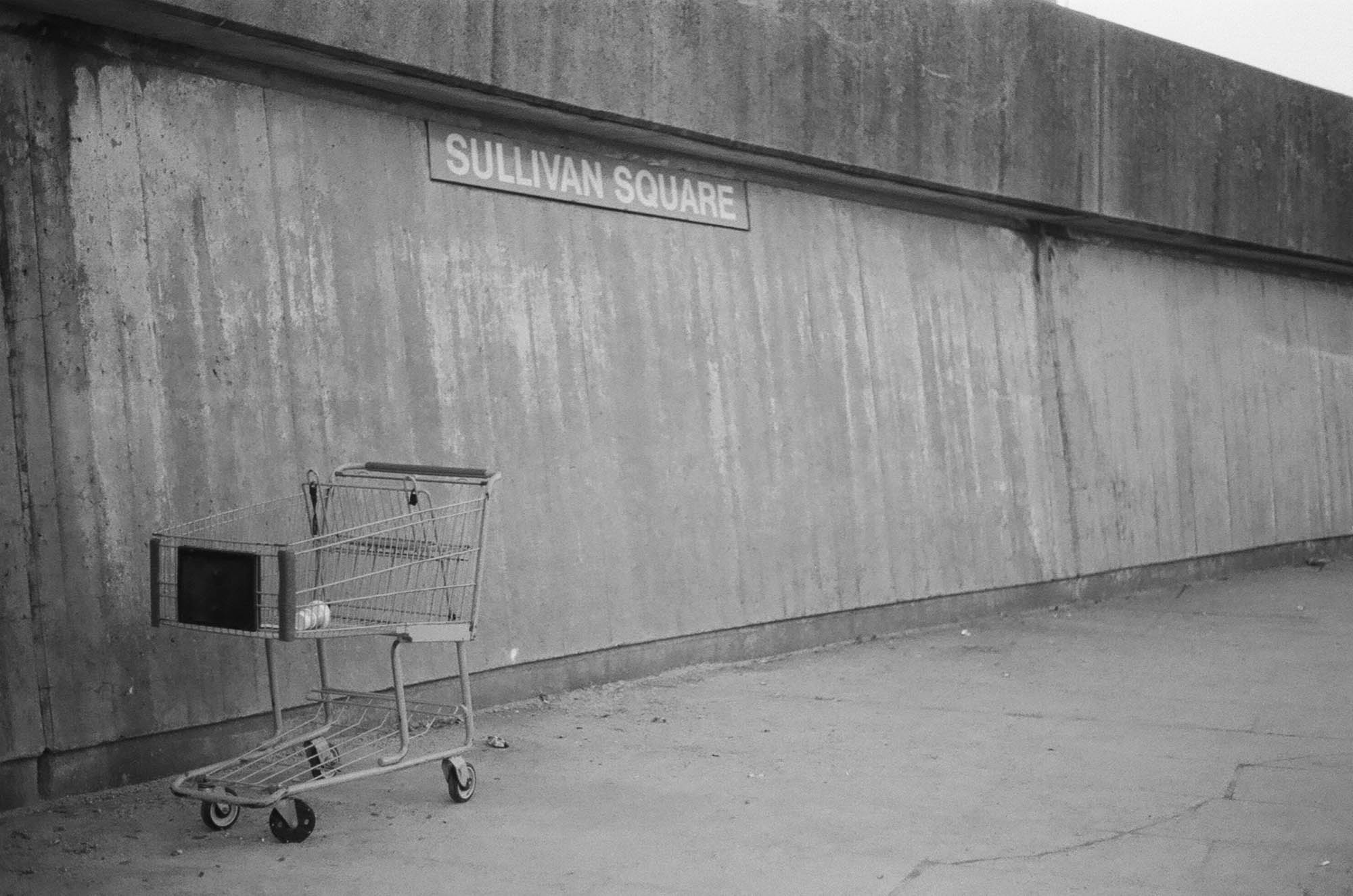 A concrete wall outside the Sullivan Square orange line stop with a shopping cart in the foreground