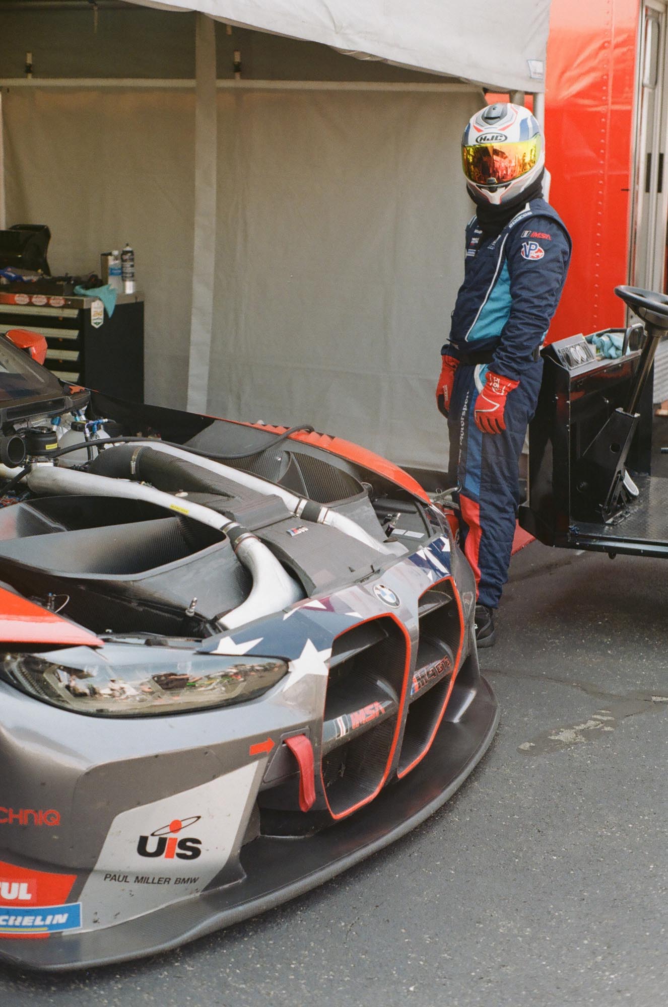 A driver with their helmet on looking at the camera, near their BMW race car